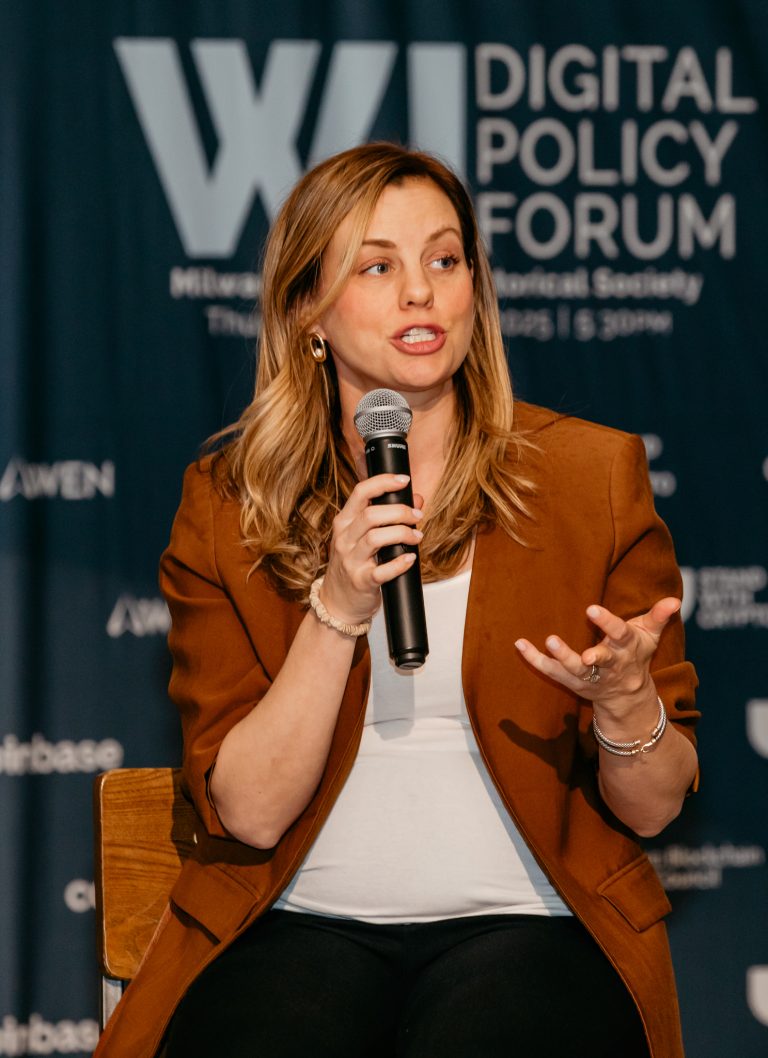 A speaker with a microphone in front of the Wisconsin Digital Policy Forum backdrop, mid-sentence.